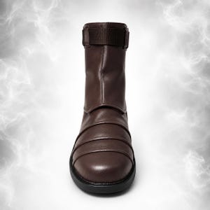 May include: A dark brown mid-calf boot featuring a smooth, rounded toe and a series of horizontal, layered panels across the vamp. The upper shaft includes a matching brown fabric strap with a buckle closure at the top, set on a black sole.