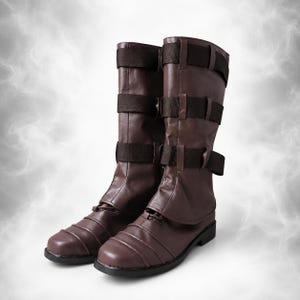 May include: A pair of mid-calf brown boots featuring three horizontal dark brown fabric straps with buckles across the shaft. The boots have a rounded toe, a flat sole, and a layered design on the lower foot section with visible stitching details.