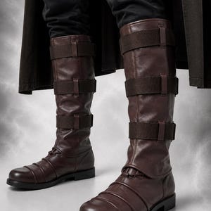 May include: A pair of dark brown knee-high boots featuring three horizontal fabric straps across the calf and a series of stitched horizontal ridges across the rounded toe area. The boots have a flat black sole and a smooth, matte finish.