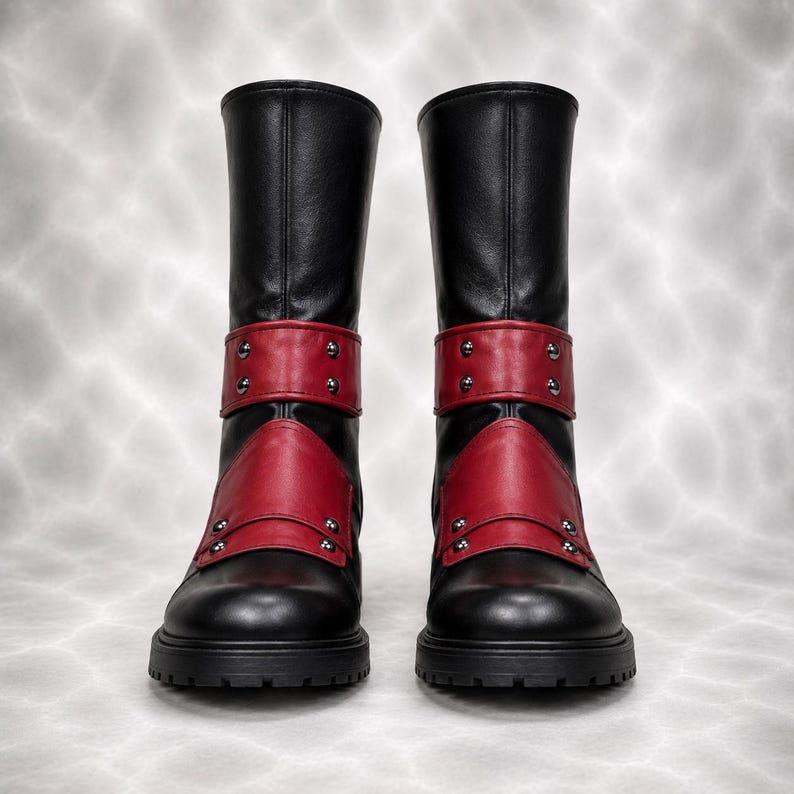 May include: A pair of mid-calf boots in black with red accents. The boots feature a red strap across the ankle and a red panel over the instep, both secured with silver-toned metal studs. The boots have a rounded toe and a thick, lugged black sole.