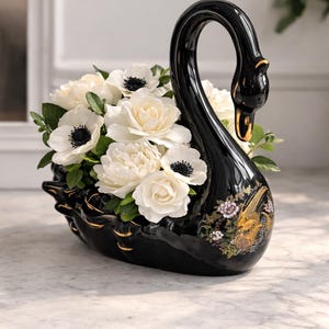 Puede incluir: Un jarrón negro de cerámica con forma de cisne lleno de flores blancas y vegetación. El jarrón tiene detalles dorados y un diseño decorativo floral y de pájaros. Las flores incluyen anémonas blancas con centros negros y rosas blancas.