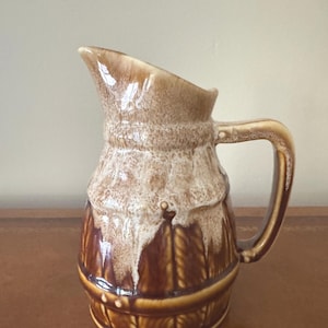 Puede incluir: Una jarra de cerámica con un esmalte marrón y beige. La jarra tiene un asa y un pico para verter. El diseño incluye un efecto texturizado y en capas con una banda decorativa en la base. La jarra probablemente se utiliza para servir bebidas.