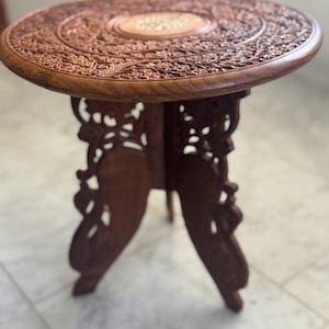 Puede incluir: Una mesa auxiliar redonda de madera marrón oscuro con intrincadas tallas florales en la mesa. La mesa tiene tres patas ornamentadas. La mesa tiene aproximadamente 50 cm de diámetro y 50 cm de alto.