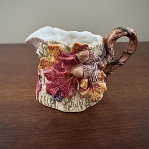 Puede incluir: Una pequeña jarra de cerámica con asa, decorada con hojas de otoño, bellotas y uvas. La jarra tiene una base texturizada en forma de corteza y un interior blanco. El asa está diseñada para parecerse a una rama de árbol.