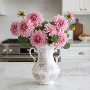 以下が含まれることがあります： ピンクのダリアと緑の葉が入った白い磁器の花瓶。花瓶には2つの取っ手があり、紫と黄色の花柄があります。花は満開で、鮮やかなディスプレイを作り出しています。花瓶は白い大理石のカウンタートップに置かれています。