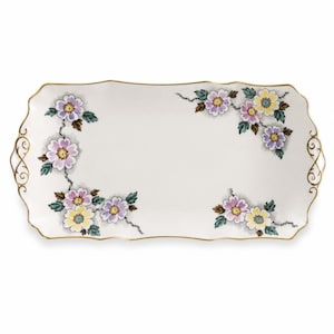 May include: A rectangular white ceramic serving tray with scalloped edges and gold trim. The tray features floral patterns in shades of purple, yellow, and green, with delicate gold filigree handles on the left and right sides of the piece.