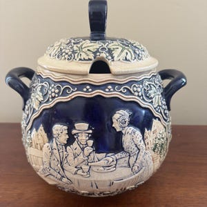 May include: A dark blue and cream ceramic lidded jar with two side handles. The exterior features a raised relief scene of three people at a table, surrounded by embossed grapevines and leaves. The lid includes a notch for a spoon or ladle.