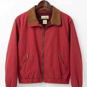 May include: A red jacket with a brown suede collar and a full zipper closure. The jacket has two front pockets with zippers and ribbed cuffs and hem. The jacket is hanging on a wooden hanger.