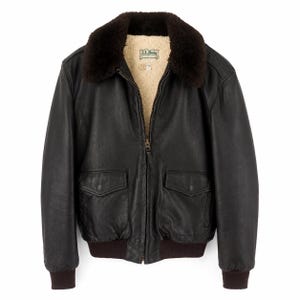 May include: A black leather bomber jacket featuring a dark brown faux fur collar, a cream-colored fleece lining, and a front zipper closure. The jacket includes two front flap pockets, ribbed cuffs, and a ribbed hem, with an L.L.Bean label inside.