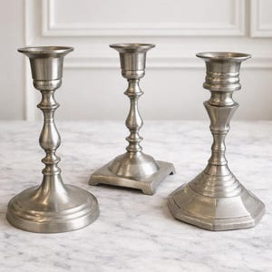 May include: Three silver-toned metal candlesticks with varying base and stem designs. The candlesticks are displayed on a marble surface, showcasing their elegant and classic aesthetic. The candle holders are suitable for home decor.