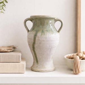 Puede incluir: Un jarrón de cerámica con dos asas, cuerpo cónico y borde ancho. La superficie tiene un acabado esmaltado con un color verde salvia en la parte superior que gotea hacia una base de color blanco crema, creando un aspecto texturizado.