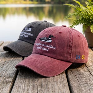 Può includere: Due cappellini da baseball con testo ricamato e grafica di uccello. Un cappellino è grigio scuro, l'altro bordeaux. Il cappellino bordeaux ha il testo "stupid canadian wolf bird" e i numeri "24 81". Entrambi i cappellini sono su una superficie di legno.