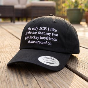 May include: Black adjustable baseball cap with white text: "the only ICE I like is the ice that my two gay hockey boyfriends skate around on." The cap has a curved brim and a silver tag.