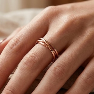 Könnte beinhalten: Ein Ring aus Roségold mit einem mehrreihigen Design aus drei dünnen, ineinander verschlungenen Metallsträngen. Der Ring wird an einem Finger getragen und weist eine polierte, glatte Oberfläche auf, die das Licht reflektiert.