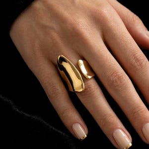 Peut inclure: Une bague dorée au design ouvert et asymétrique, composée de deux plaques métalliques lisses, polies et aux formes organiques qui entourent le doigt. La bague présente une surface hautement réfléchissante et une structure large et audacieuse.