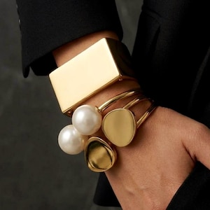 Colección de pulseras de oro y plata pulidas con diseños de doble perla ovalada y brazalete cuadrado en acero inoxidable.