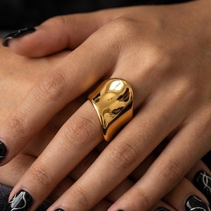 Può includere: Un anello grande, color oro, con una superficie liscia e riflettente. L'anello ha un design curvo unico. La mano ha lo smalto nero con disegni a onde argentate. Un gioiello audace.