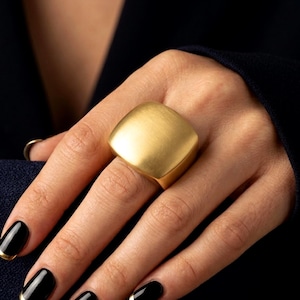 Könnte beinhalten: Ein großer, klobiger Goldring mit einer glatten, matten, quadratischen Oberfläche und abgerundeten Kanten. Der Ring hat ein massives, solides Banddesign, das markant auf dem Finger sitzt und ein sauberes, poliertes Metallfinish aufweist.