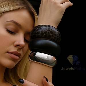 Può includere: Un set di tre bracciali sovrapposti indossati al polso. Il bracciale superiore è marrone scuro con un motivo testurizzato e maculato. Il bracciale centrale è nero opaco, liscio e uniforme. Il bracciale inferiore è in metallo argentato lucido.