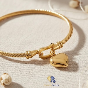 Può includere: Un bracciale rigido color oro in stile cavo con un ciondolo a forma di cuore lucido sospeso a una chiusura a gancio. Il gioiello presenta una texture metallica intrecciata lungo il cerchio e una finitura liscia e riflettente sul cuore.