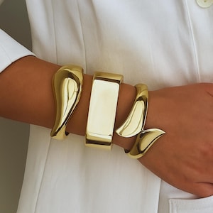 Può includere: Tre bracciali in metallo color oro indossati al polso. Il set comprende un bracciale rigido liscio e curvo, una fascia rettangolare larga e lucida e un bracciale avvolgente con estremità affusolate. I gioielli hanno una finitura metallica lucida.