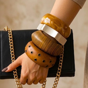Può includere: Una borsa rettangolare nera con tracolla a catena dorata e quattro bracciali larghi sovrapposti. I bracciali includono un pezzo in resina ambrata marmorizzata, una fascia liscia color argento, un polsino effetto legno e una fascia color cuoio con occhielli.