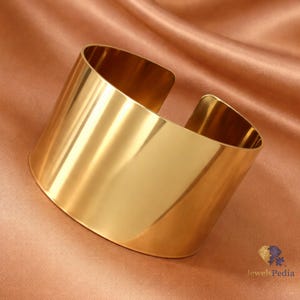 Può includere: Un ampio bracciale a fascia color oro lucido con una superficie liscia e riflettente e un design aperto. Il bracciale presenta una struttura solida e minimalista con una finitura lucida che cattura la luce, creando dei riflessi brillanti.