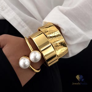 Può includere: Set di gioielli color oro con un paio di anelli con perle e tre braccialetti larghi e strutturati. I braccialetti hanno una trama martellata e una finitura liscia e lucida. I gioielli sono indossati sopra una camicia bianca e abiti neri.