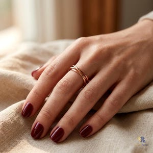 May include: A rose gold ring featuring a multi-band design with three thin, intertwined metallic strands. The ring is worn on the ring finger, showcasing a polished, smooth finish that reflects light across its circular, layered structure.