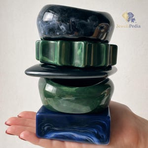 May include: A stack of colorful resin bangles in shades of blue and green. The top bangle is a dark blue with white swirls, followed by a green, scalloped bangle. The stack is held in a hand.