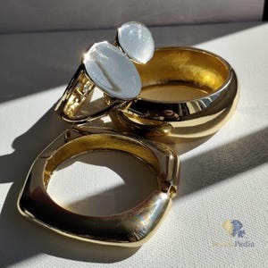 May include: A selection of gold-coloured jewellery, including two rings with oval, iridescent stones and two gold bangles. The bangles have a rounded and a square design. The jewellery is displayed on a white surface, with sunlight creating shadows.