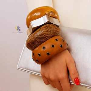 Può includere: Una pila di braccialetti in legno e metallo nei toni del marrone e dell'argento. I braccialetti includono un braccialetto grande e arrotondato in legno, un pezzo geometrico argentato, un braccialetto in legno a righe e un braccialetto in pelle marrone con occhielli metallici. La mano tiene una pochette argentata.