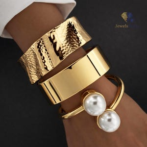 Può includere: Bracciali dorati. L'immagine presenta tre bracciali dorati: un bracciale a polsino strutturato, una fascia larga e liscia e un bracciale curvo con due grandi perle bianche. I bracciali sono indossati su un polso, su sfondo scuro.