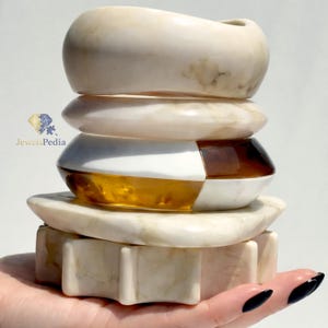 Può includere: Una pila di braccialetti decorativi in pietra e una base a forma di stella. I braccialetti sono nelle tonalità crema, bianco e marrone. La base è color crema con un design a forma di stella. L'immagine include il testo "JewelsPedia".