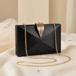 Può includere: Una pochette rettangolare nera con superficie testurizzata e design geometrico sfaccettato. La borsa presenta una chiusura in metallo dorato a forma di foglia nella parte superiore centrale e include una sottile tracolla a catena dorata.