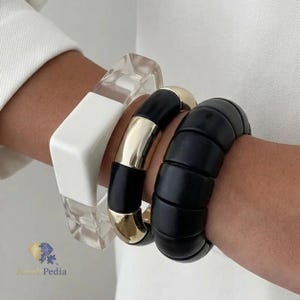 Può includere: Un set di tre bracciali composto da un bracciale in acrilico trasparente e bianco, un bracciale in metallo nero e color oro e un ampio bracciale nero segmentato. I bracciali sono indossati insieme, mostrando un mix di texture lisce e lucide.