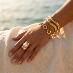 Puede incluir: Un anillo dorado con una gran perla blanca central, acompañado de dos brazaletes dorados gruesos y ondulados, y una pulsera de cadena delicada compuesta por pequeños eslabones circulares. El conjunto es de metal dorado pulido.
