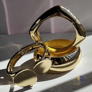 Può includere: Sono presentati tre braccialetti color oro. Uno ha una forma quadrata, un altro una forma arrotondata e il terzo è un braccialetto a cerniera con due accenti circolari. I gioielli sono riflettenti e poggiano su una superficie bianca.