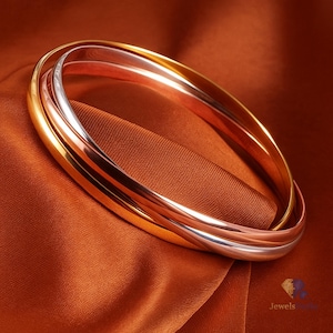 Può includere: Tre braccialetti a cerchio intrecciati in oro, oro rosa e argento. Le fasce lisce e lucide risaltano su uno sfondo di tessuto color ruggine. I gioielli sono esposti in un ambiente da studio, evidenziando la lucentezza metallica dei braccialetti.