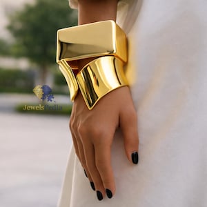 Könnte beinhalten: Ein goldfarbenes Statement-Armband mit rechteckiger Oberseite und geschwungenem, offenem Design. Das Armband ist hochglanzpoliert und reflektiert das Licht, wodurch ein kühner und moderner Look entsteht. Der Schmuck wird am Handgelenk getragen.