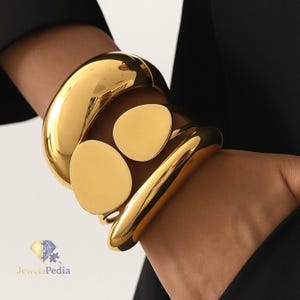 May include: A gold-coloured bracelet with a polished finish. The bracelet features a thick, rounded band and three flat, circular accents. The jewellery is worn on a wrist, with a black garment in the background. The brand name "Jewels Pedia" is visible.