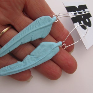 May include: A pair of light blue feather earrings with silver hooks. The earrings are made of polymer clay and are shaped like feathers.