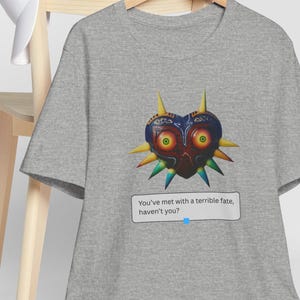 Könnte beinhalten: Ein graues T-Shirt mit einem farbenfrohen Majora's Mask-Motiv. Das Design zeigt eine herzförmige Maske mit gelben Stacheln und dem Text: "You've met with a terrible fate, haven't you?" Eine weiße Baseballkappe liegt auf einem Holzstuhl.