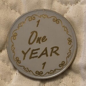 May include: A silver-colored commemorative coin with gold lettering and accents. The coin reads "1 One YEAR 1" in a stylized font. The border features a decorative gold filigree design with hearts and swirls.