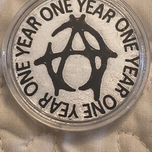 May include: A silver coin encased in a clear plastic holder. The coin features the words "ONE YEAR" in a circular pattern around a black stylized "A" symbol. The coin has a shiny, reflective surface.