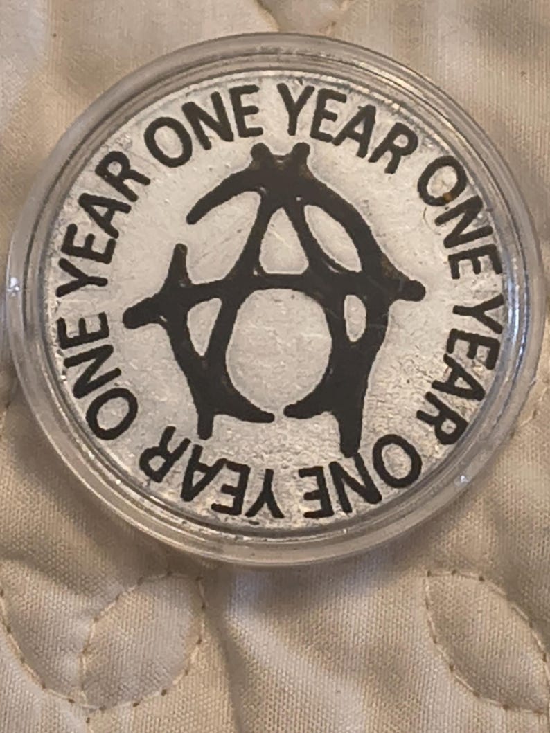 May include: A silver coin encased in a clear plastic holder. The coin features the words "ONE YEAR" in a circular pattern around a black stylized "A" symbol. The coin has a shiny, reflective surface.