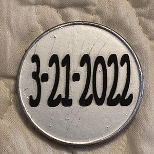 Puede incluir: Un objeto redondo, de color plateado, con la fecha "3-21-2022" impresa en negrita, en negro. El objeto tiene un borde met&aacute;lico y parece ligeramente rayado. El fondo es una tela acolchada de color blanquecino.