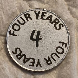 Puede incluir: Una ficha met&aacute;lica plateada con las palabras "FOUR YEARS" en un patr&oacute;n circular en el borde. El n&uacute;mero "4" est&aacute; en el centro. La ficha tiene un aspecto ligeramente desgastado.