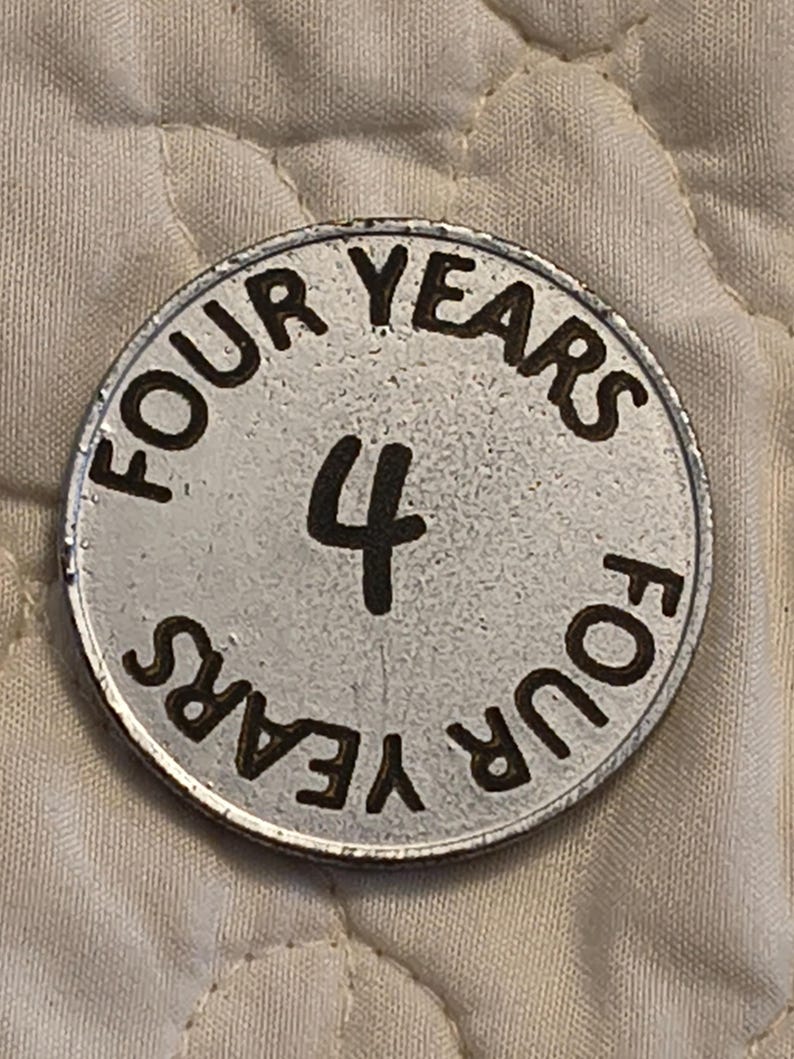 Puede incluir: Una ficha met&aacute;lica plateada con las palabras "FOUR YEARS" en un patr&oacute;n circular en el borde. El n&uacute;mero "4" est&aacute; en el centro. La ficha tiene un aspecto ligeramente desgastado.