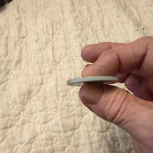May include: A silver coin held between fingers, showing the edge of the coin. The coin has a reeded edge and appears to be a quarter. The background is a cream-colored, quilted fabric.